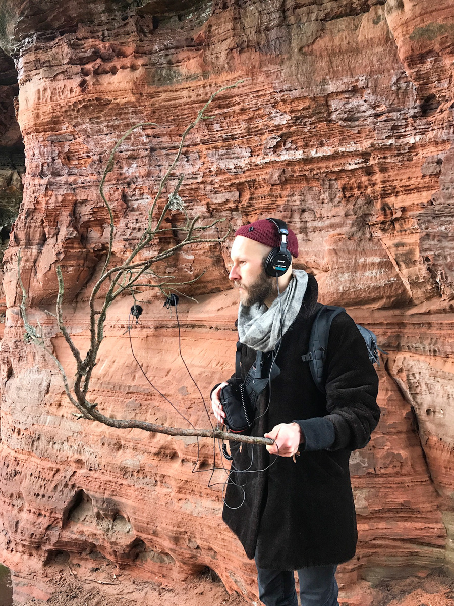 Timm Roller holding a branch of a tree with microphones attached, outside in nature.